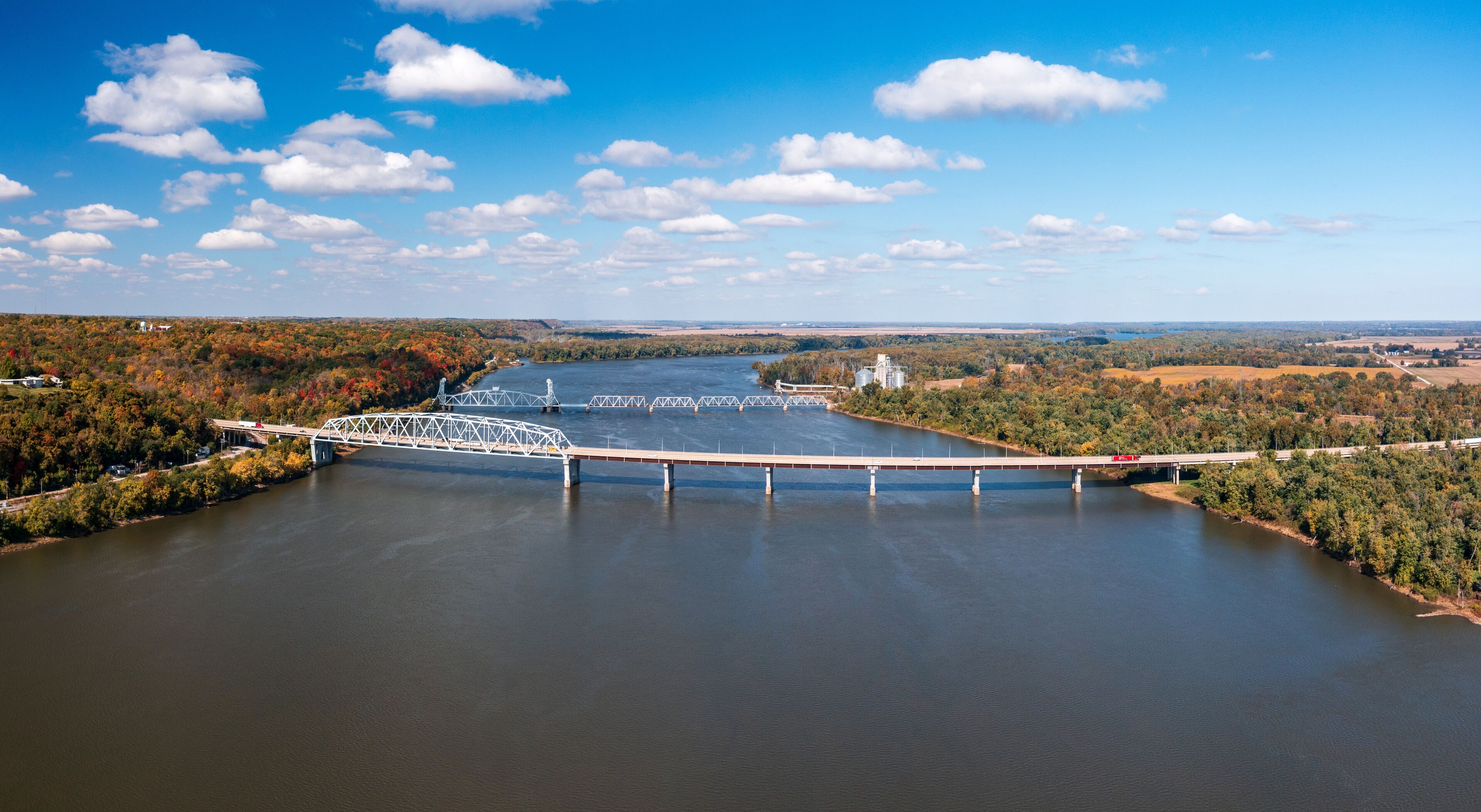 A bridge over a river with autumn foliage and industrial structures in the background.