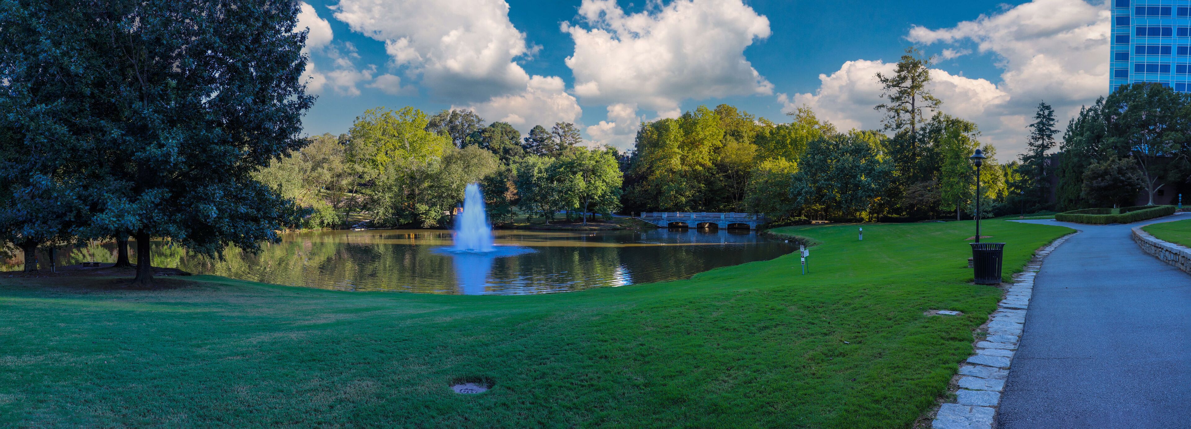 a stunning panoramic shot of a lake with a waterfall in the middle of the water surrounded by lush green trees and grass with blue sky and clouds at Lenox Park in Brookhaven Georgia USA