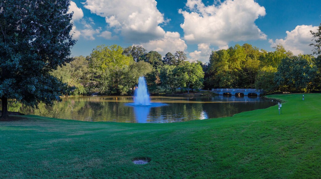 a stunning panoramic shot of a lake with a waterfall in the middle of the water surrounded by lush green trees and grass with blue sky and clouds at Lenox Park in Brookhaven Georgia USA