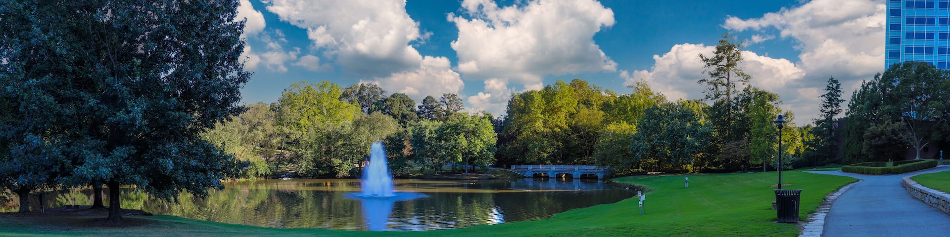 a stunning panoramic shot of a lake with a waterfall in the middle of the water surrounded by lush green trees and grass with blue sky and clouds at Lenox Park in Brookhaven Georgia USA