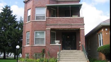 A two-story brick building with a balcony, a front door, and steps leading up to the entrance.