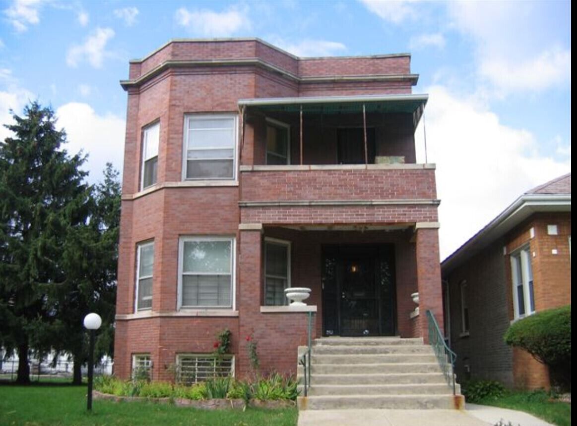 A two-story brick building with a balcony, a front door, and steps leading up to the entrance.