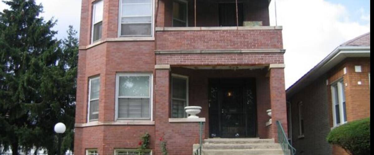 A two-story brick building with a balcony, a front door, and steps leading up to the entrance.