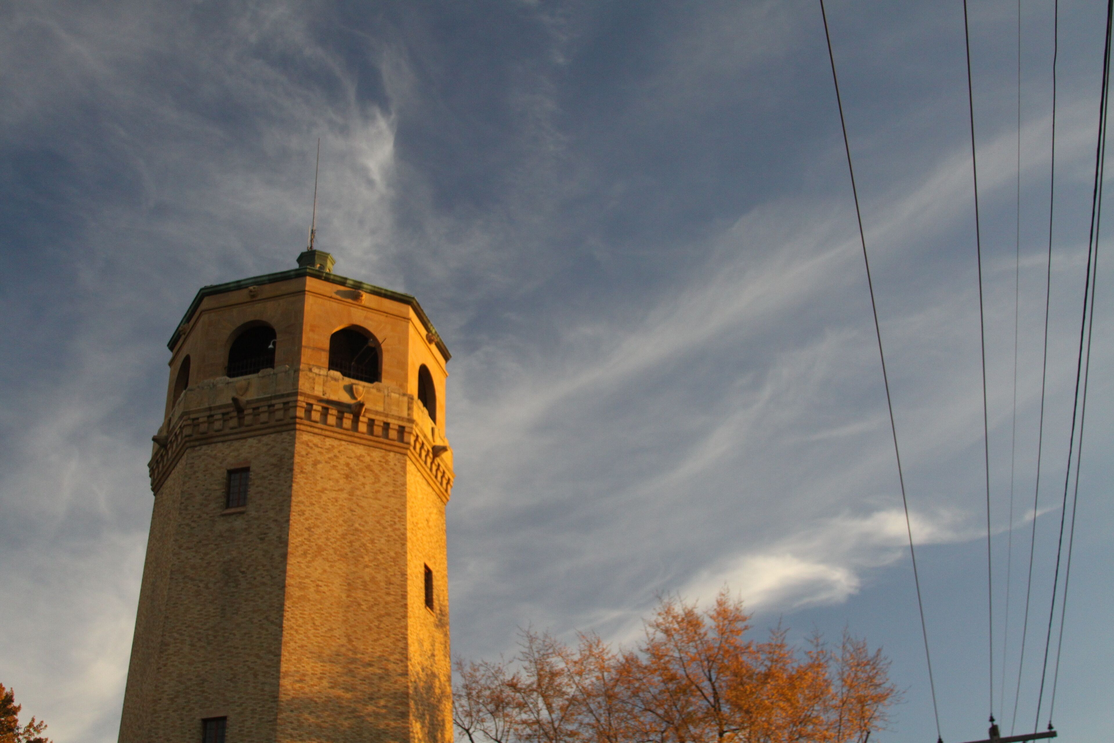 A tall tower with arched windows and a clock, surrounded by autumn trees and power lines against a blue sky.