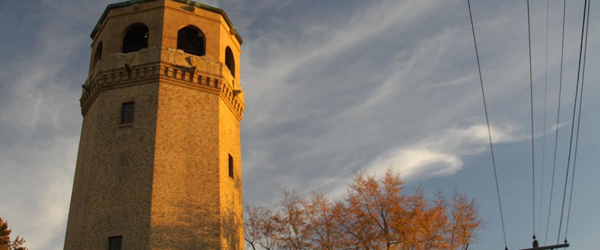 A tall tower with arched windows and a clock, surrounded by autumn trees and power lines against a blue sky.