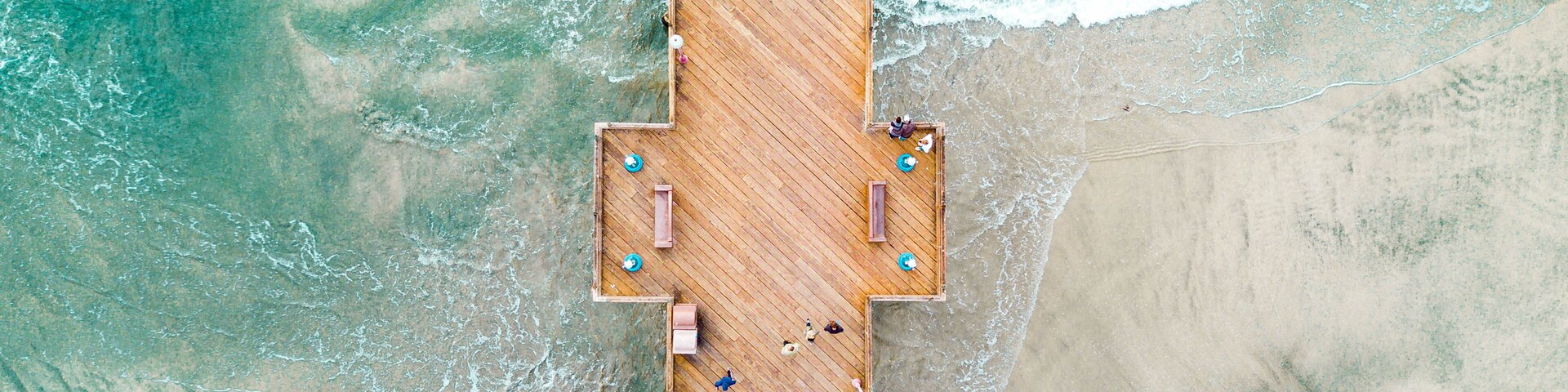 Vista aérea de un muelle de madera que se extiende hacia aguas turquesas, con gente caminando por él.