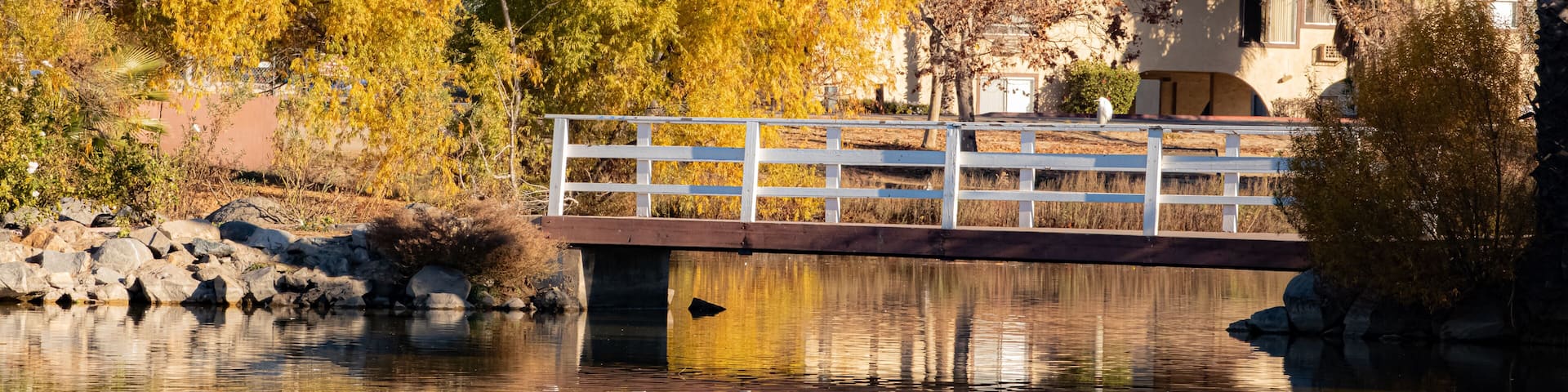 A bridge over a calm river with autumn-colored trees reflecting in the water, surrounded by hills.