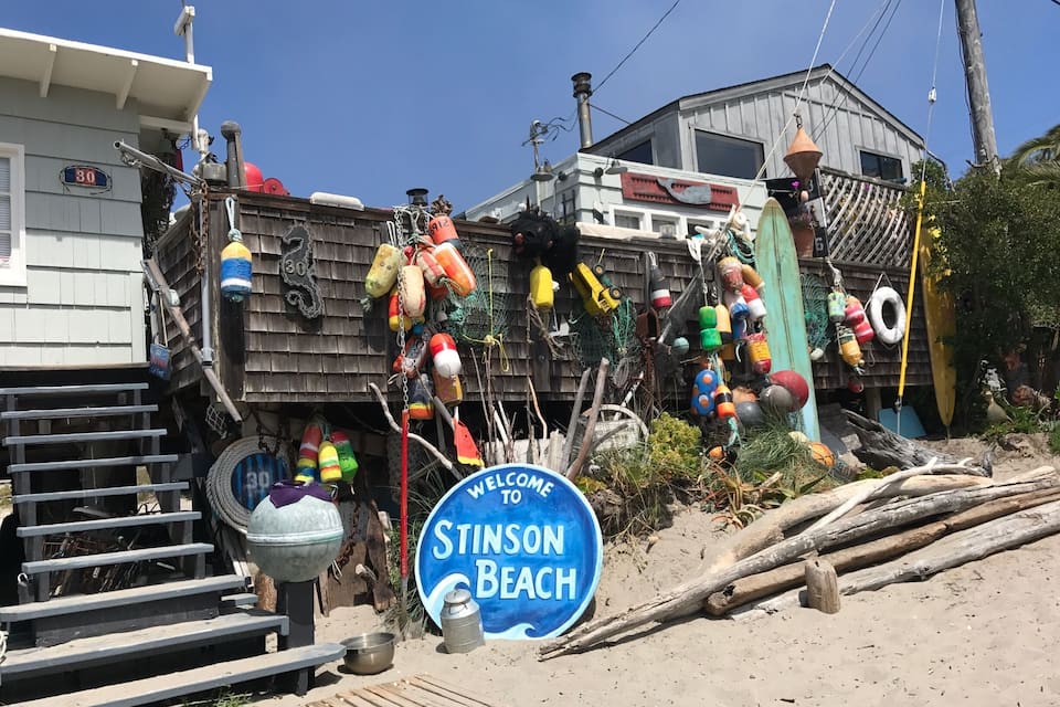 A cluttered outdoor area with a 'Welcome to Stinson Beach' sign, various colorful buoys, and a wooden structure.