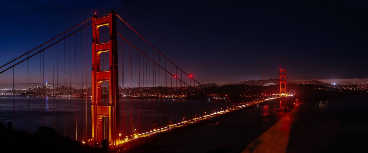 A nighttime view of the Golden Gate Bridge with its iconic red towers and cables, spanning across a body of water with city lights in the background.