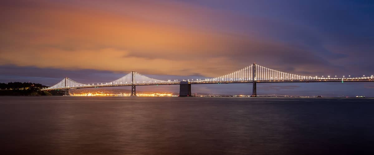 A long-span suspension bridge at night with illuminated cables, spanning a wide river.