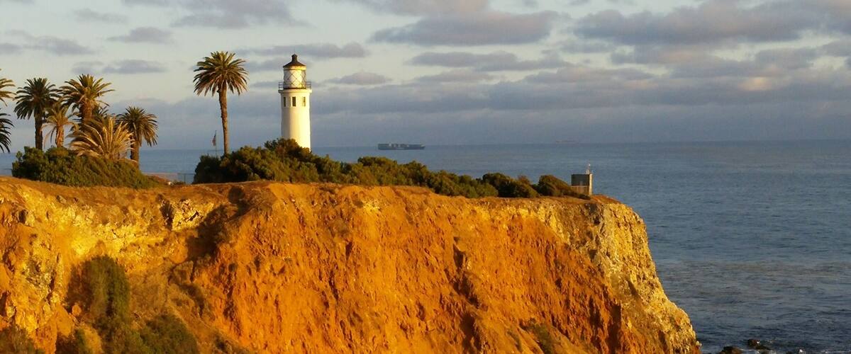 A coastal landscape with a lighthouse, palm trees, and a rocky cliff overlooking the sea.