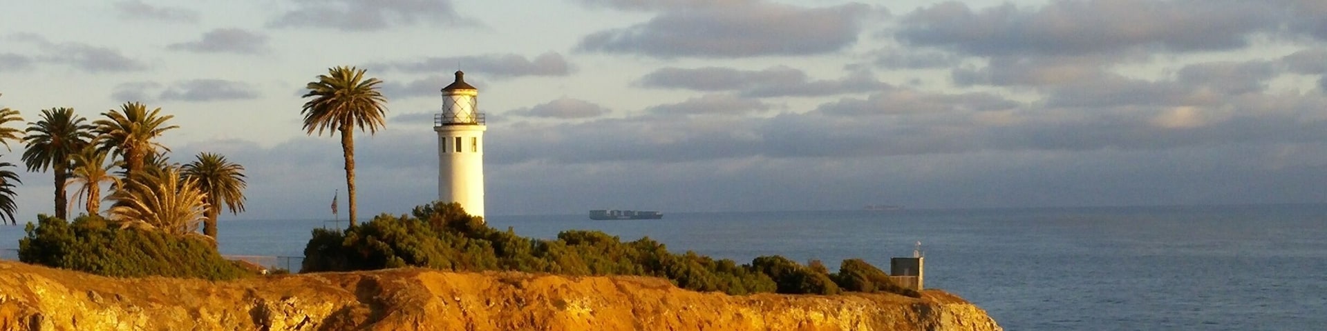 A coastal landscape with a lighthouse, palm trees, and a rocky cliff overlooking the sea.
