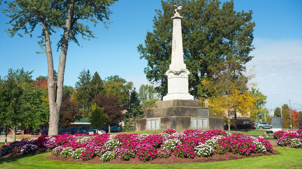 A white monument with an eagle on top, surrounded by a flower bed with pink and white flowers, and trees in the background.