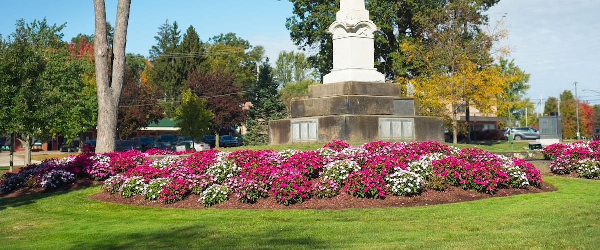 A white monument with an eagle on top, surrounded by a flower bed with pink and white flowers, and trees in the background.