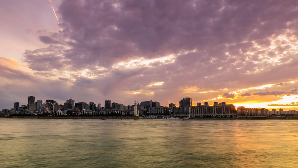 A city skyline at sunset with a prominent Ferris wheel and a body of water in the foreground.