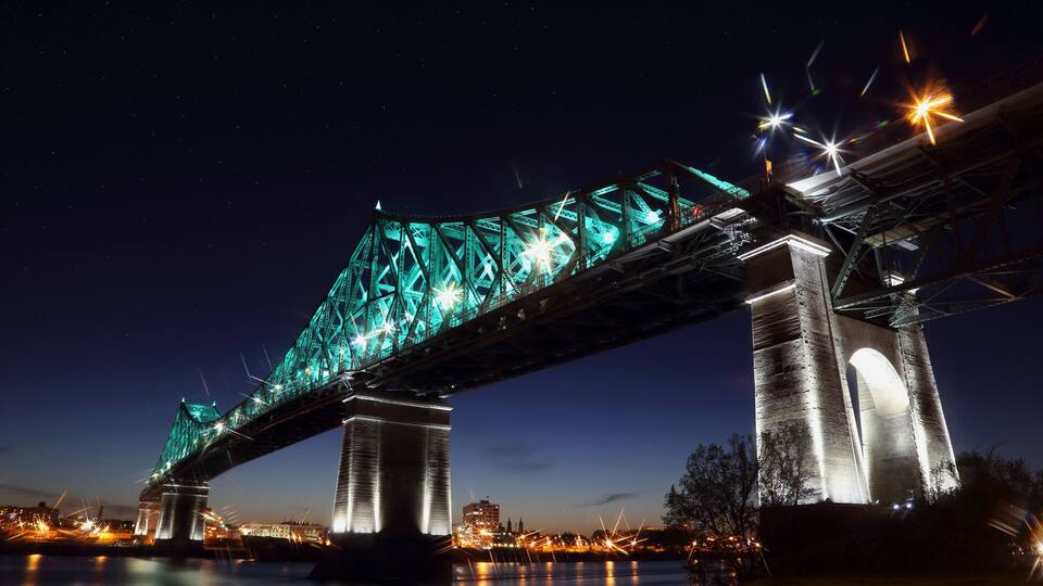 A nighttime view of a large bridge with green lighting, spanning over a body of water with city lights in the background.