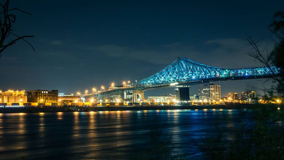 A nighttime view of a large bridge with blue lights, a river, and city buildings in the background.