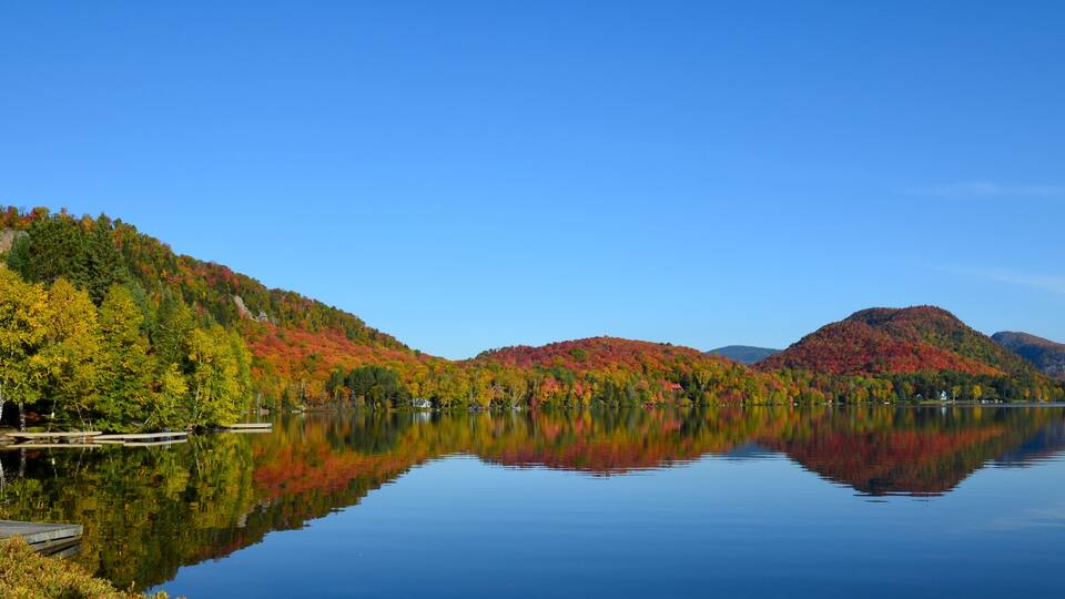 A calm lake with reflections of autumn-colored trees and distant mountains.