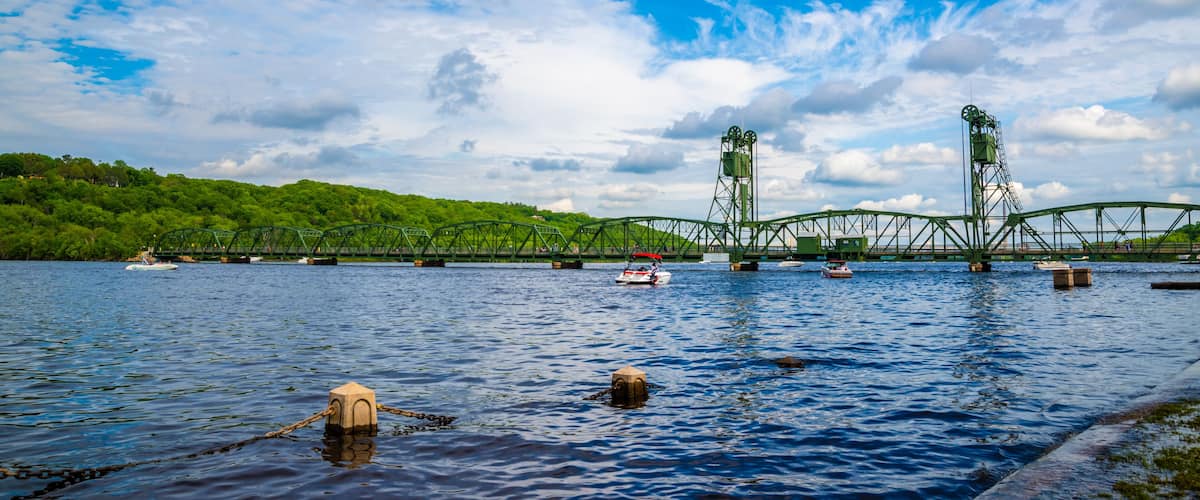 A green bridge over a river with boats and greenery in the background.