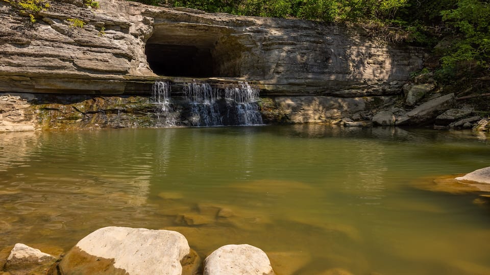 A waterfall cascading into a clear pool surrounded by large rocks and a rocky cave entrance.