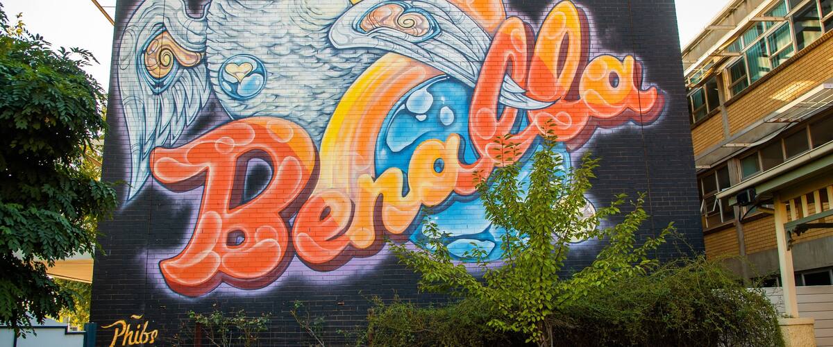 Un mural con un gran texto colorido que dice 'Benalla' y un diseño estilizado de un ave sobre un muro de ladrillos.
