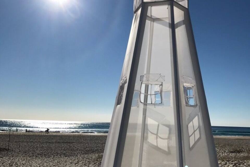 A tall, transparent lighthouse on a beach with the sun shining in the background.