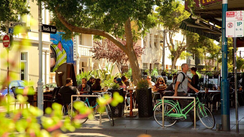 Rua movimentada com pessoas sentadas em cafés ao ar livre, bicicletas estacionadas e um letreiro indicando 'back bar'.