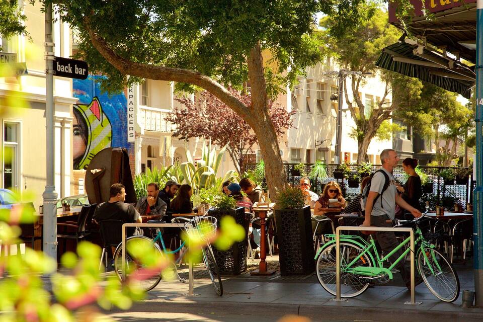A busy street with people sitting at outdoor cafes, bicycles parked, and a 'back bar' sign.
