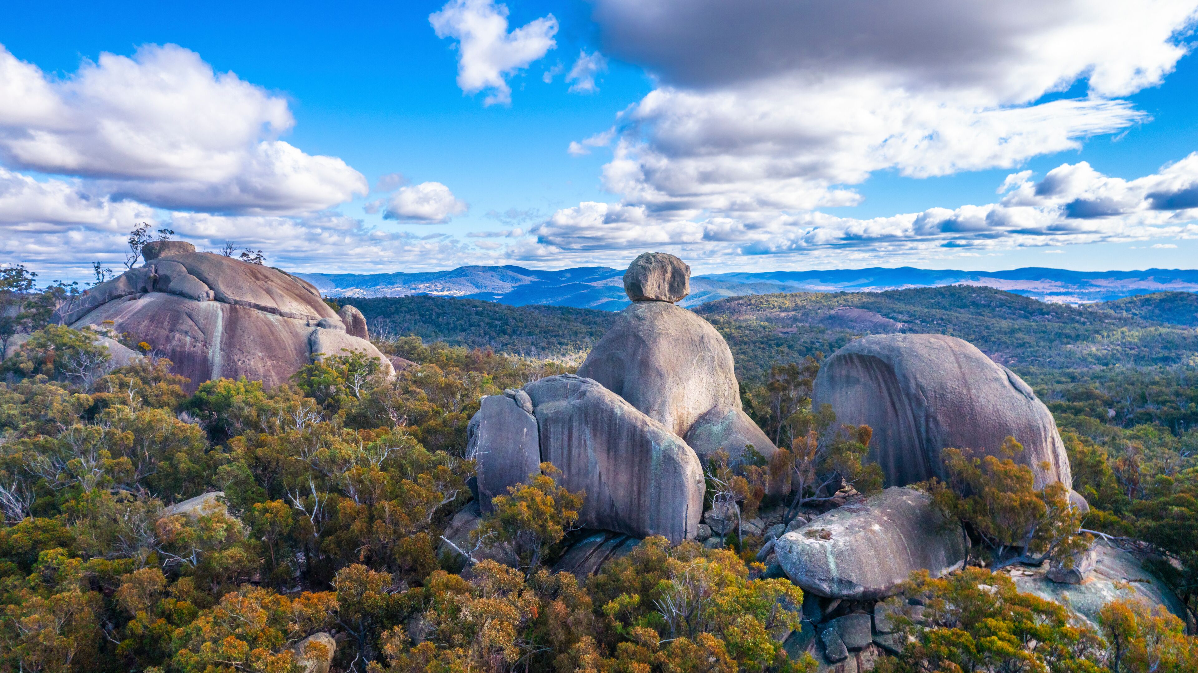 A landscape with large rock formations, dense forest, and distant mountains under a partly cloudy sky.