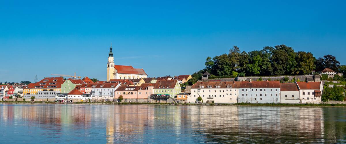 A riverside town with colorful buildings, a prominent church tower, and a castle on a hill.
