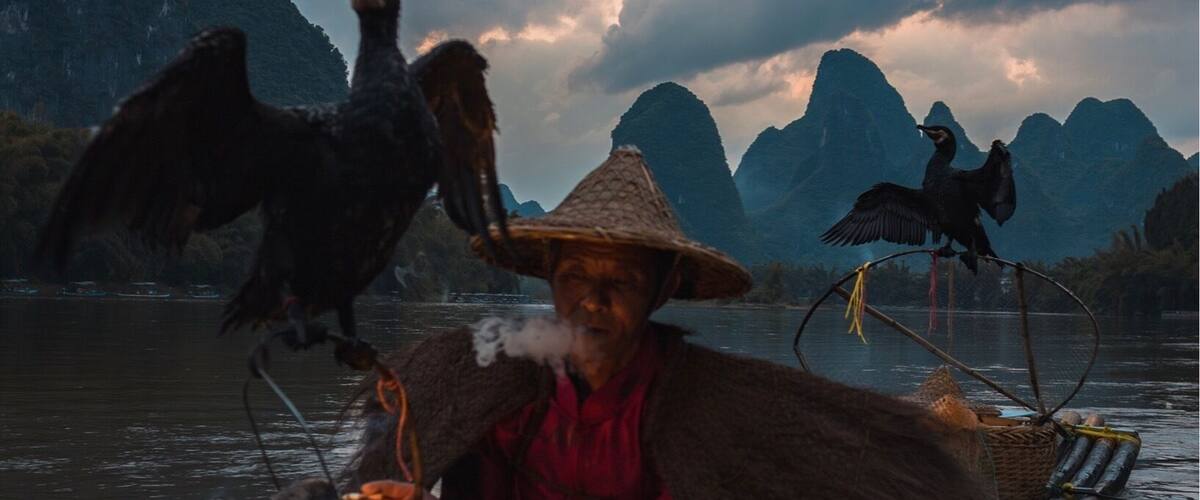 A person in a traditional boat with a lantern, surrounded by mountains and birds.
