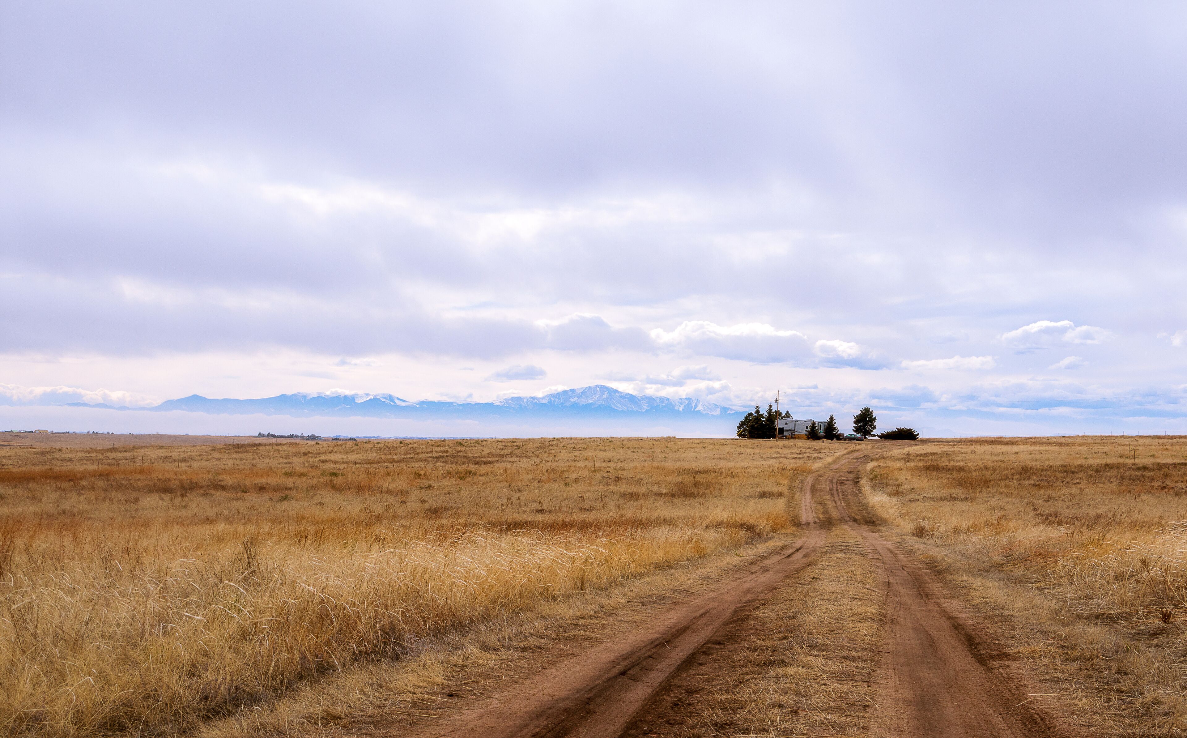 A dirt road leading to a remote house in a grassy field with mountains in the background.