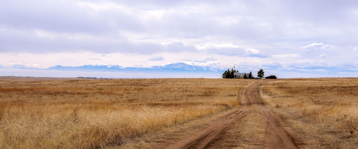 A dirt road leading to a remote house in a grassy field with mountains in the background.