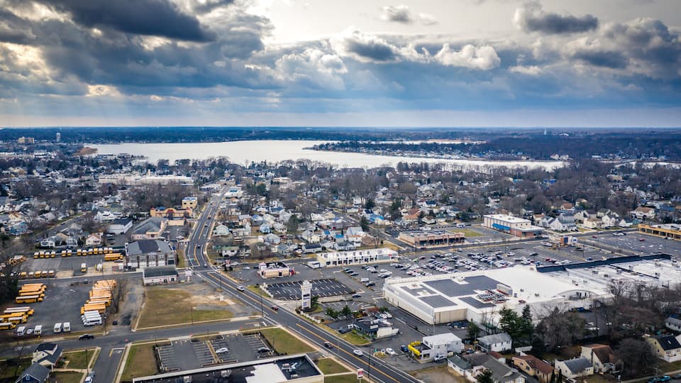 An aerial view of a town with a large body of water in the background, a highway, and various buildings including a large warehouse.