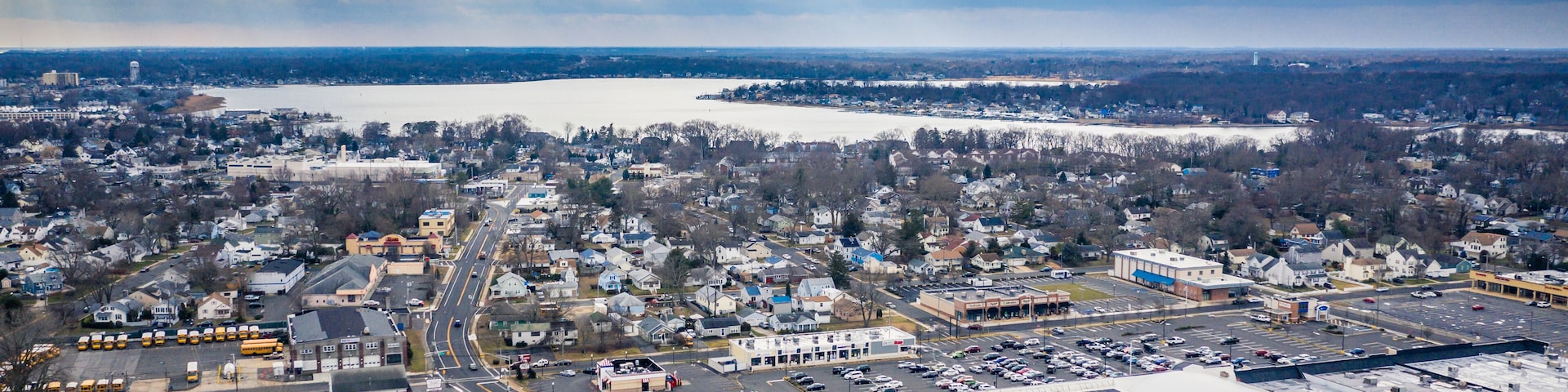 An aerial view of a town with a large body of water in the background, a highway, and various buildings including a large warehouse.