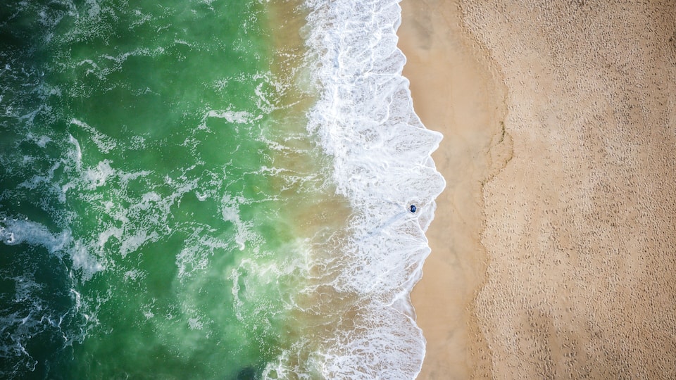 An aerial view of a beach with waves crashing onto the shore and a person standing near the water's edge.