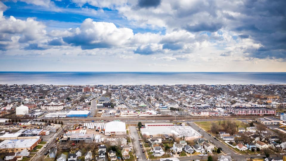 An aerial view of a coastal city with a mix of residential and industrial areas, a large body of water in the distance, and a partly cloudy sky.