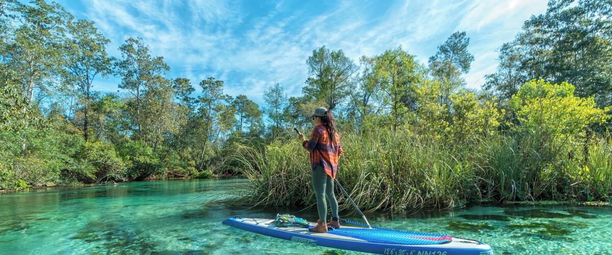 A person paddleboarding on a clear river surrounded by dense vegetation and trees.
