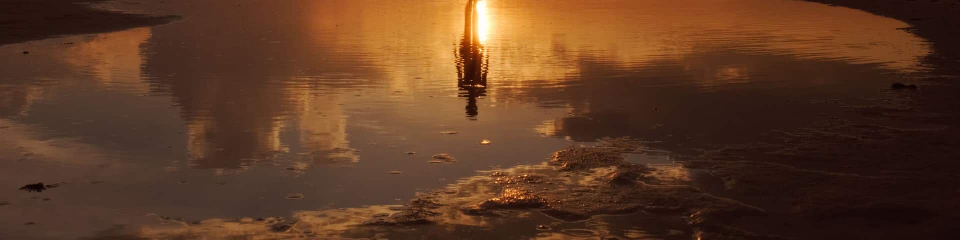 A person standing on a reflective beach at sunset with clouds in the sky.