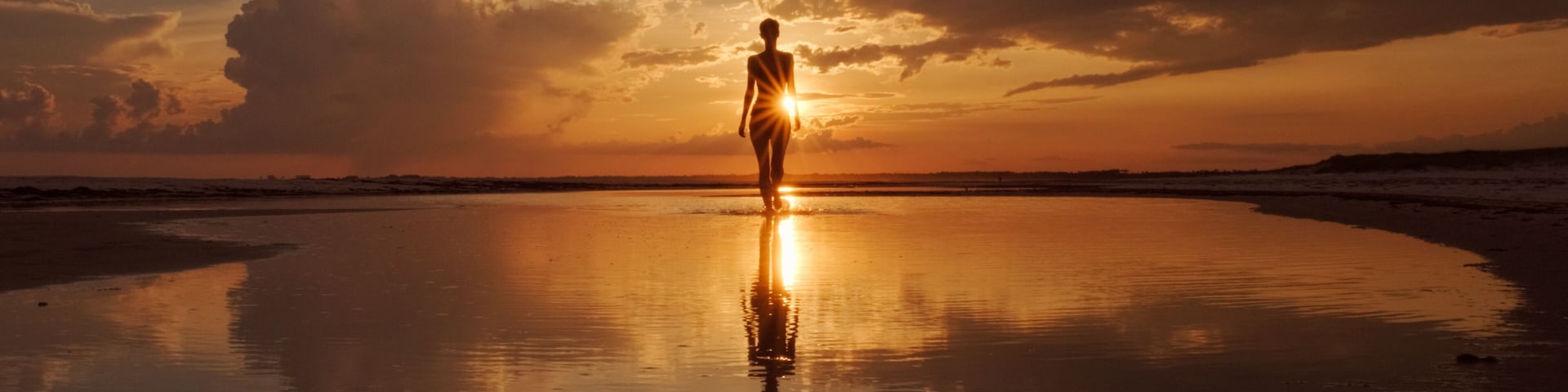 Uma pessoa em pé numa praia refletora ao pôr do sol, com nuvens no céu.