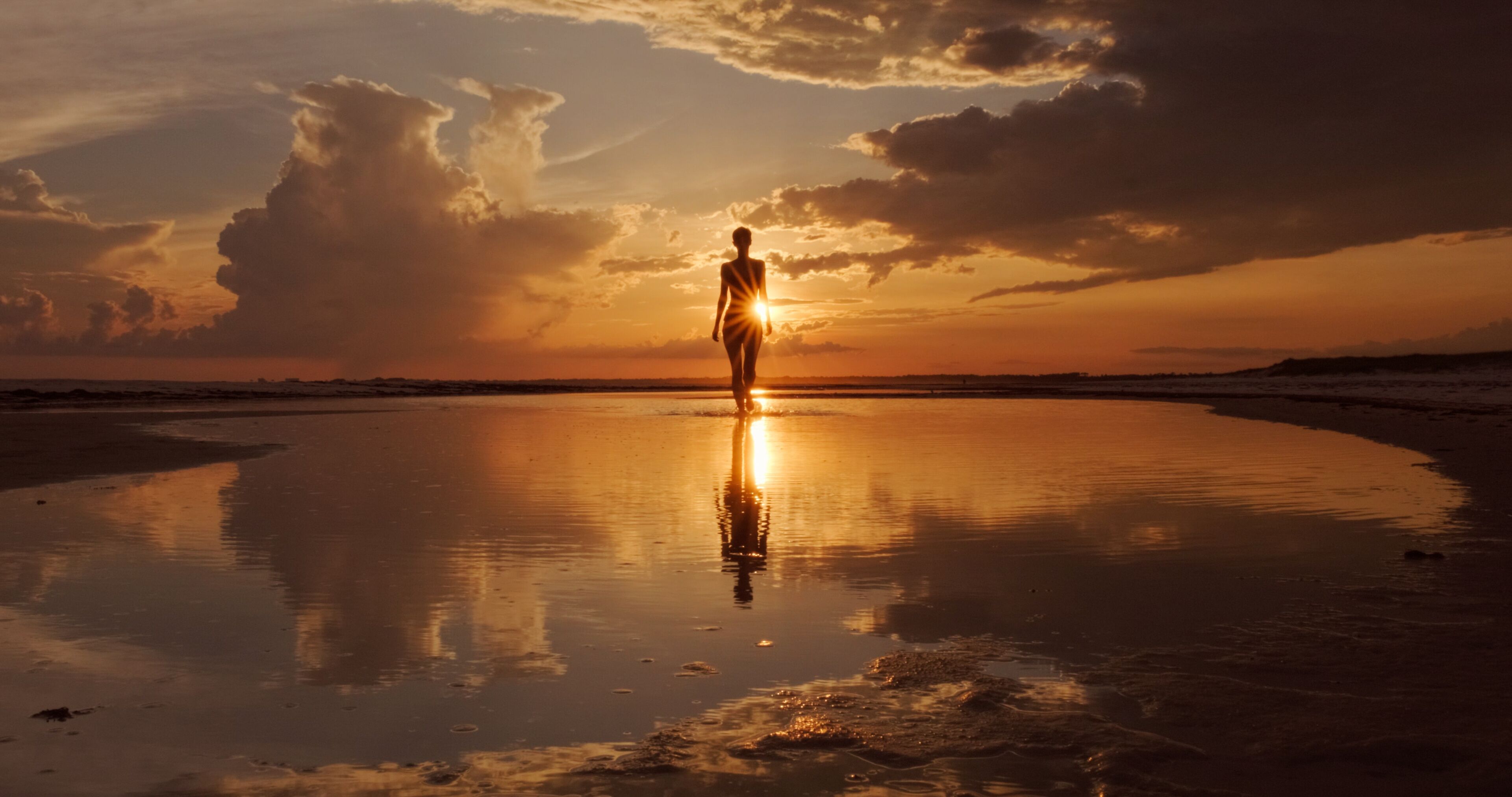 A person standing on a reflective beach at sunset with clouds in the sky.