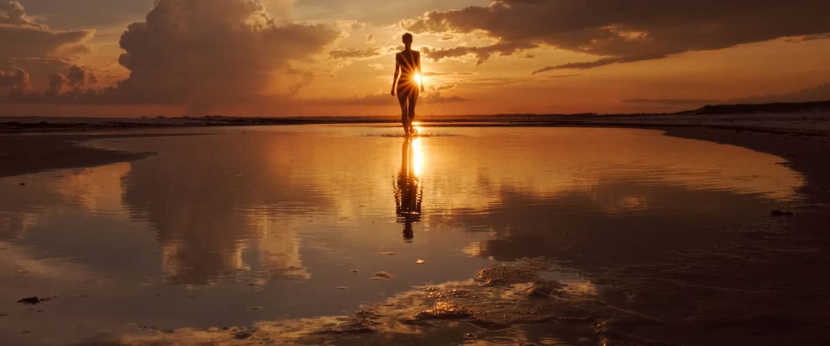 A person standing on a reflective beach at sunset with clouds in the sky.
