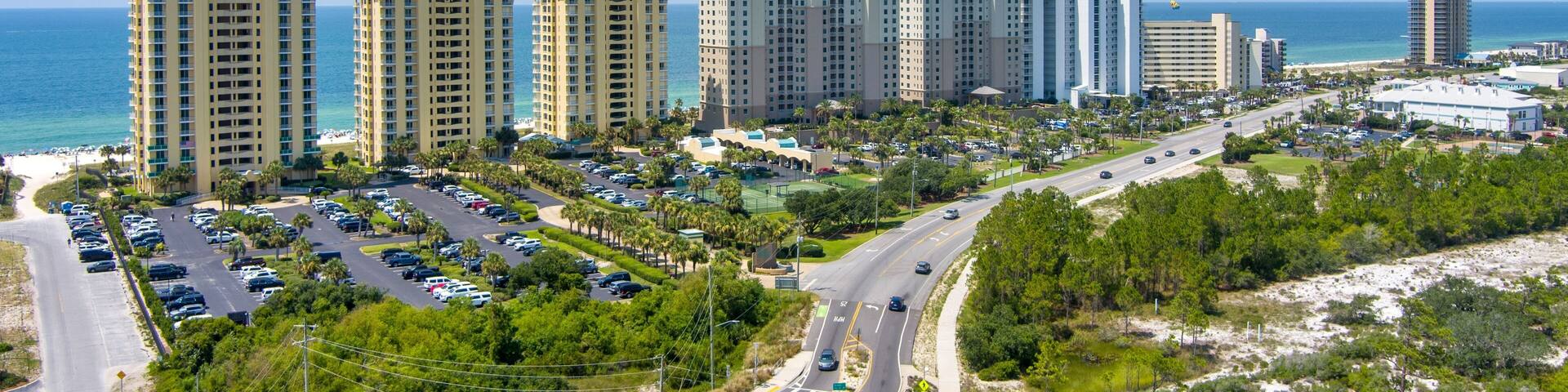 A coastal cityscape with high-rise buildings, a parking lot, and a road leading to the beach.