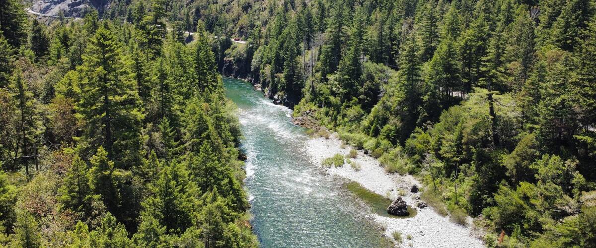Une rivière serpente à travers une vallée densément boisée, parsemée de formations rocheuses, et une route suit ses méandres le long de la rive.