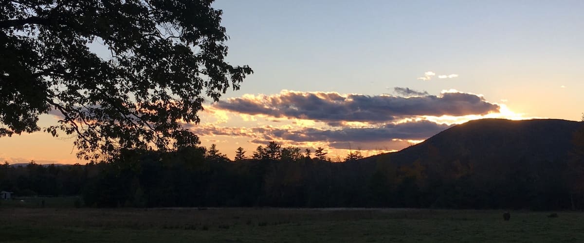 A sunset over a hilly landscape with a field in the foreground and trees silhouetted against the sky.