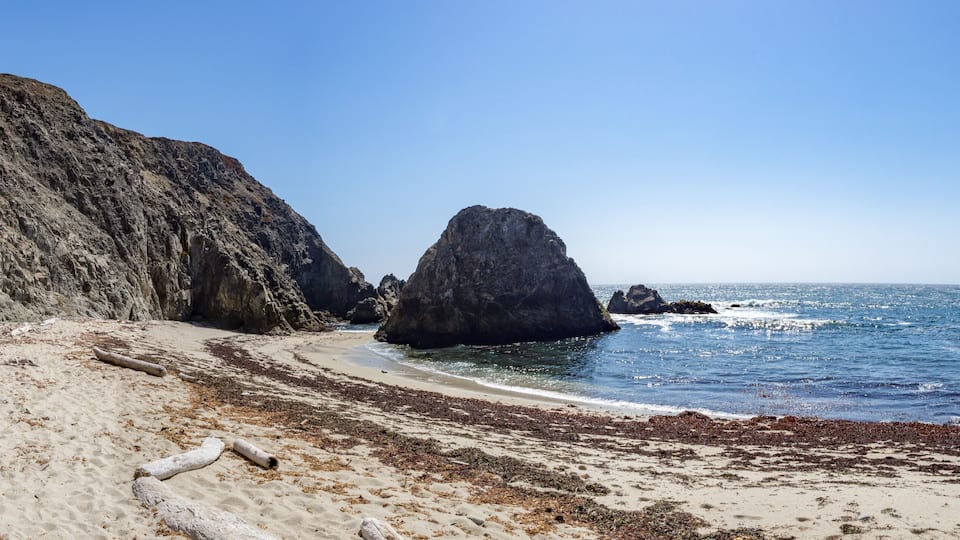 A rocky coastline with a prominent rock formation, sandy beach, and clear blue sea.