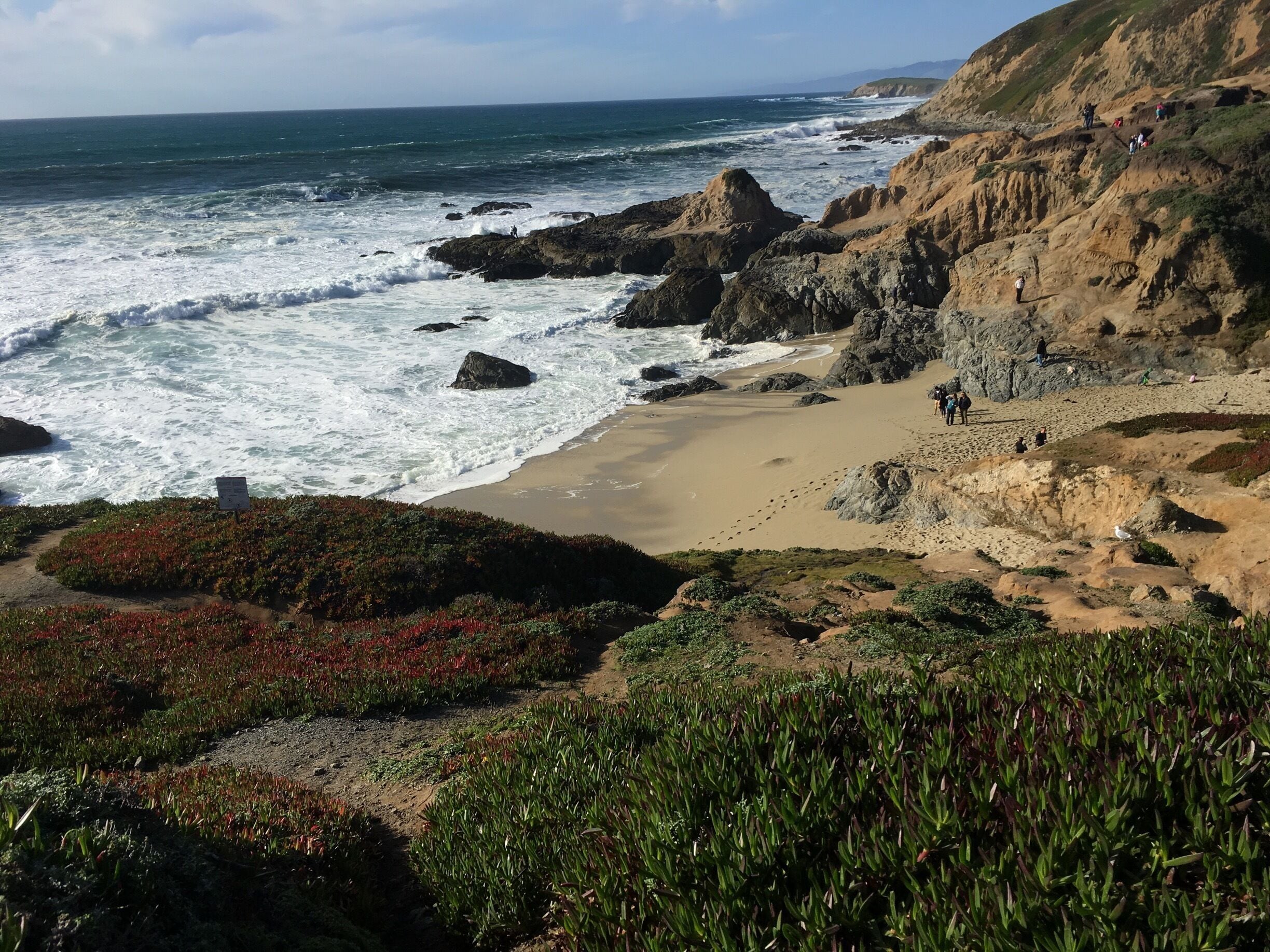 A rocky coastline with waves crashing against the shore, a sandy beach, and a trail leading down to the water.