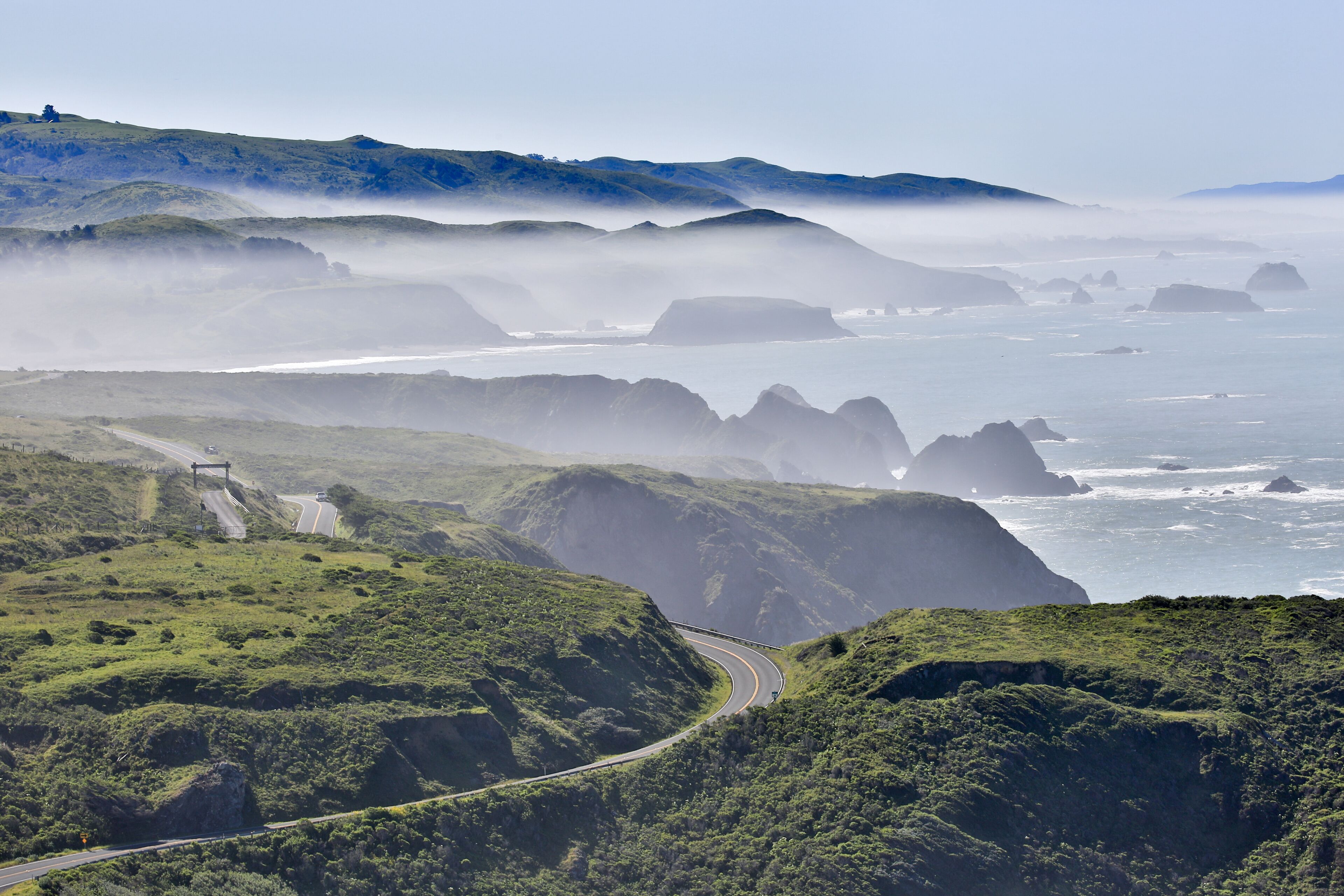 A coastal road winding through green hills with rocky outcrops and mist over the ocean.