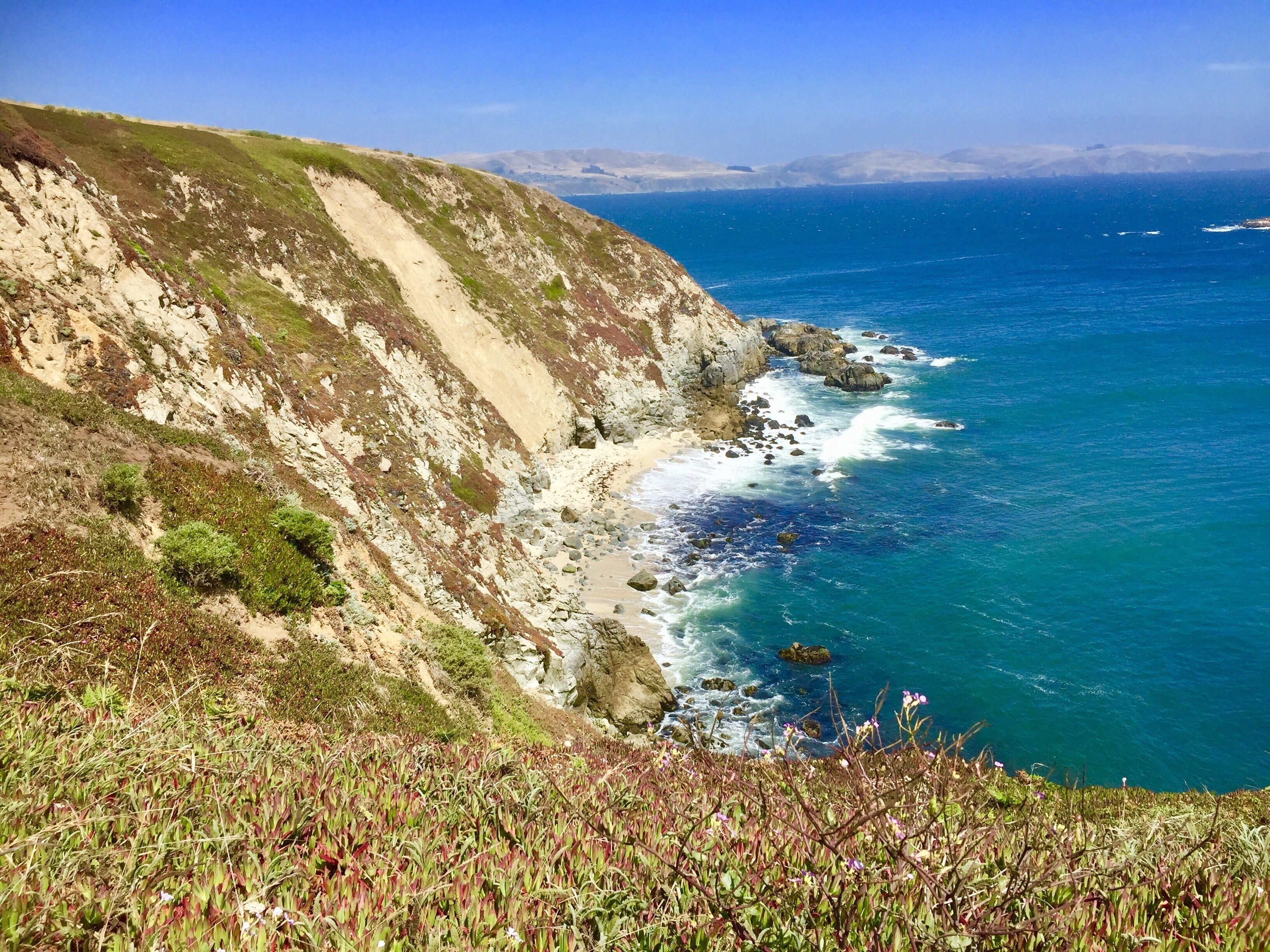 A coastal landscape with a steep cliff, rocky shoreline, and clear blue sea.