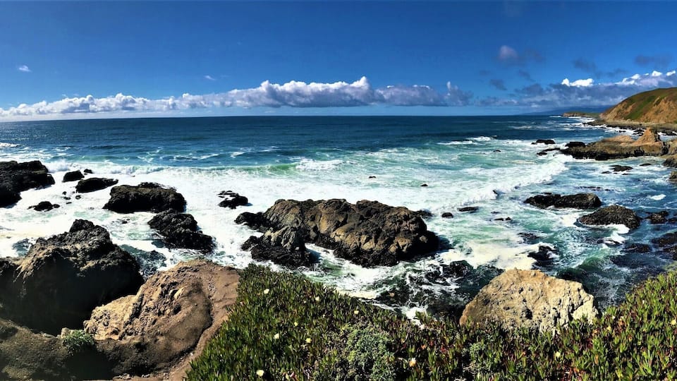 A coastal landscape with rocky cliffs, waves crashing against the shore, and a grassy hill in the background.